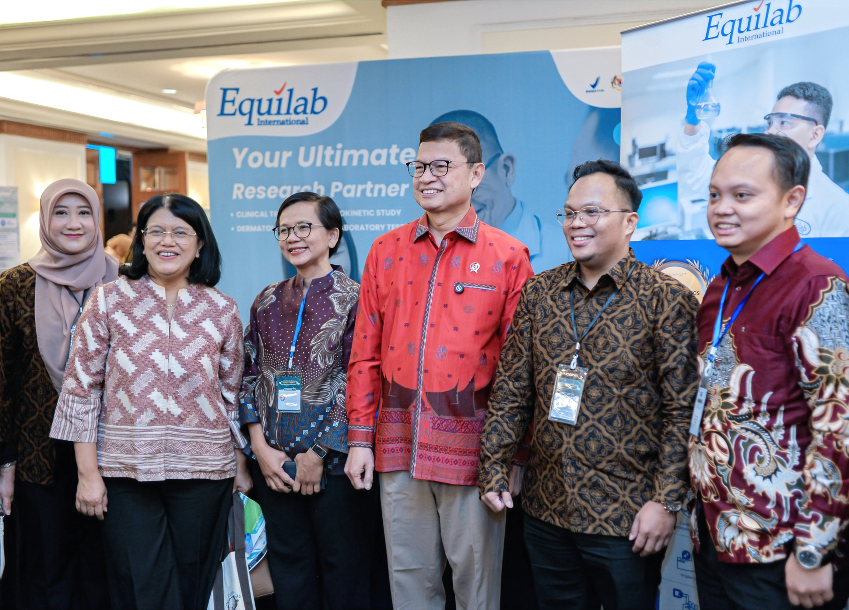 Chairperson of BPOM and five smiling adults pose at an Equilab International booth with blue banners at a conference, wearing patterned batik shirts.