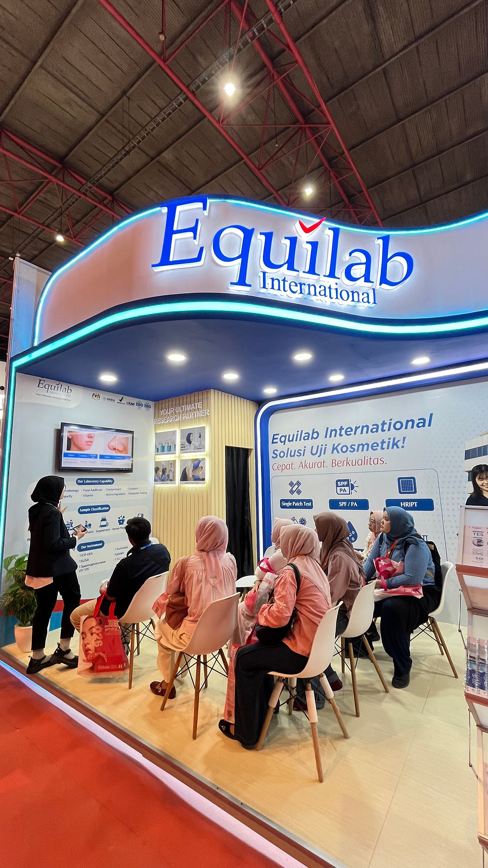 People seated at Equilab International booth, watching a presentation. Bright lighting, banners with text, and a screen on display.